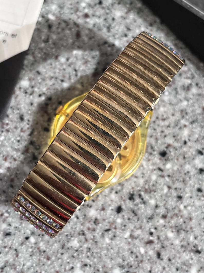 May include: Gold-tone watch with a gold-tone expandable band with clear crystals. The band has a ribbed texture.