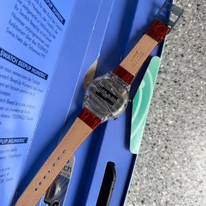 May include: A Swatch BeepUp Numeric watch with a clear face and a red leather strap is displayed in its original blue box. The watch has a rectangular face with a digital display and is accompanied by a black accessory.