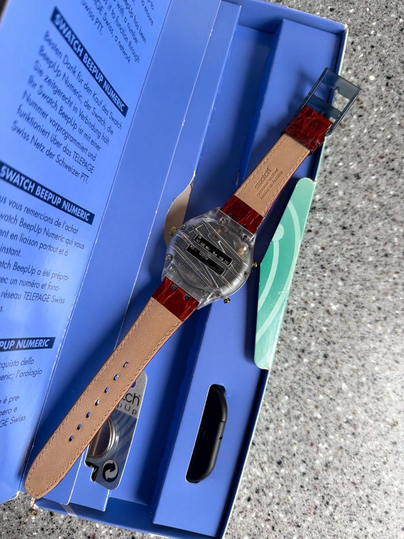 May include: A Swatch BeepUp Numeric watch with a clear face and a red leather strap is displayed in its original blue box. The watch has a rectangular face with a digital display and is accompanied by a black accessory.