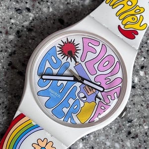 May include: White Swatch watch with a colourful, psychedelic design. The watch face features the words "Flower Power" in blue and pink, with a red sunburst and other graphics. The white strap has the word "Happy" in yellow and a rainbow.