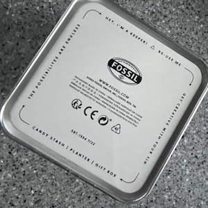 May include: A rectangular silver tin with rounded corners. The lid features the Fossil logo and text that reads "Hey, I'm a keeper! Re-use me." Additional text includes "Candy Stash | Planter | Gift Box."