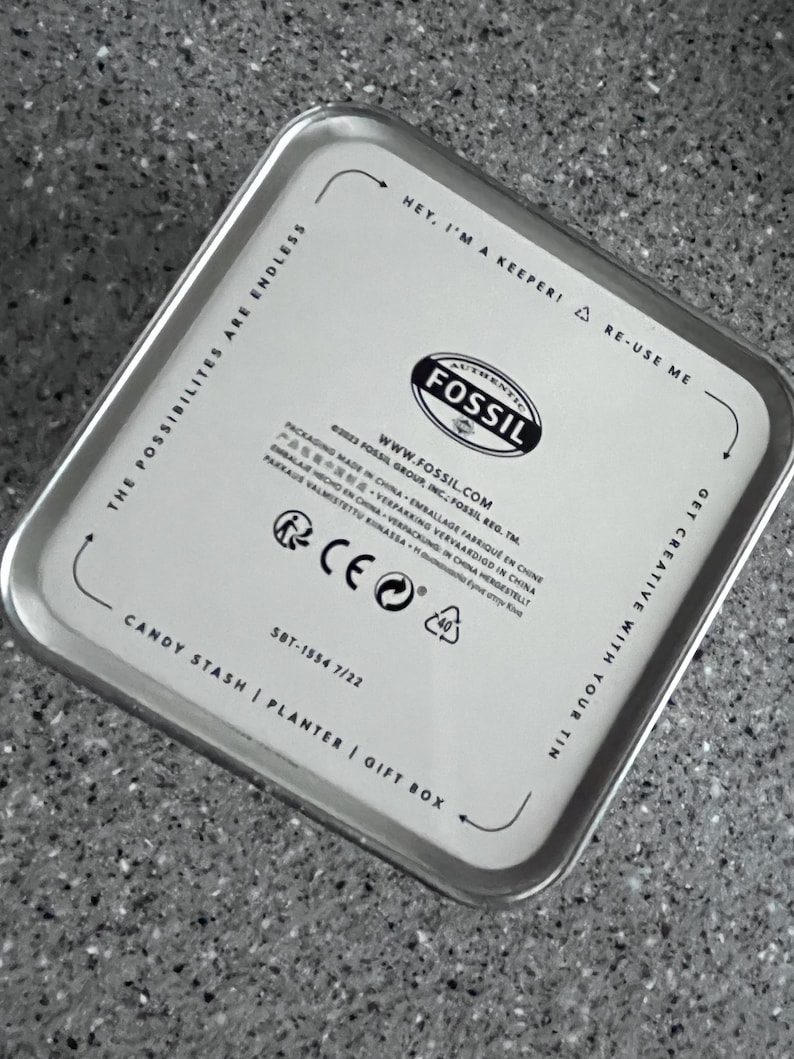 May include: A rectangular silver tin with rounded corners. The lid features the Fossil logo and text that reads "Hey, I'm a keeper! Re-use me." Additional text includes "Candy Stash | Planter | Gift Box."