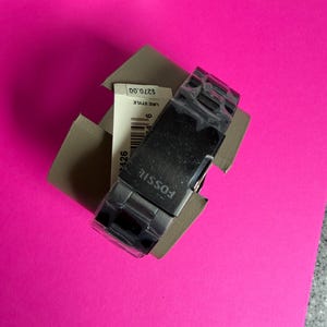 May include: A black Fossil watch with a silver band is displayed in its original packaging. The watch face is rectangular, and the band is made of interlocking links. The watch is on a bright pink surface.