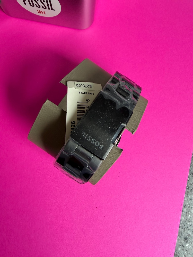 May include: A black Fossil watch with a silver band is displayed in its original packaging. The watch face is rectangular, and the band is made of interlocking links. The watch is on a bright pink surface.