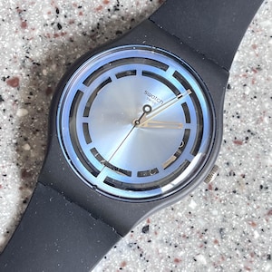 May include: A Swatch watch with a black strap and a light blue and silver face. The watch face has a circular design with the word "swatch" printed on it. The watch hands are gold.