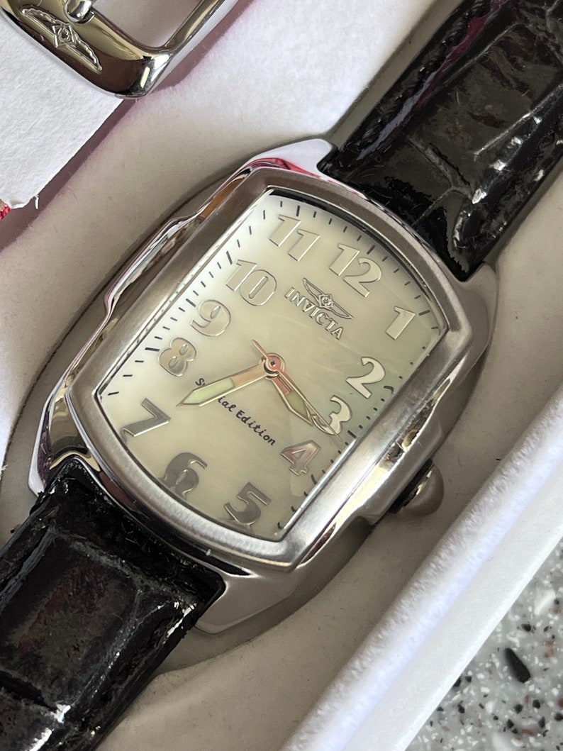 Puede incluir: Un reloj Invicta plateado con una esfera blanca y una correa de cuero negra. El reloj tiene marcadores de hora en n&uacute;meros romanos y una inscripci&oacute;n "Special Edition".
