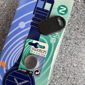 May include: A Swatch Beep Up watch battery and black watch accessory are displayed on a light blue and white patterned background. The battery is in a blister pack with the text "Swatch Beep Up". The watch accessory is black and oval-shaped.