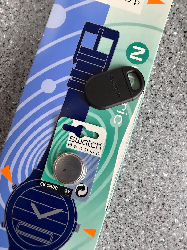 May include: A Swatch Beep Up watch battery and black watch accessory are displayed on a light blue and white patterned background. The battery is in a blister pack with the text "Swatch Beep Up". The watch accessory is black and oval-shaped.