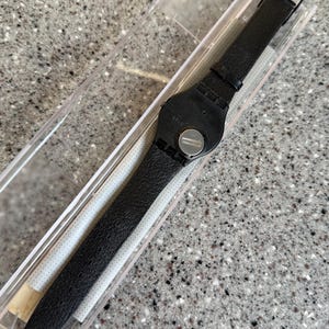 K&ouml;nnte beinhalten: Eine schwarze Swatch-Uhr mit strukturiertem Lederarmband wird in einer transparenten Kunststoffbox pr&auml;sentiert. Das Zifferblatt der Uhr ist schwarz und das Armband wird mit einer silbernen Schnalle befestigt. Die Uhr ist neu und ungetragen.