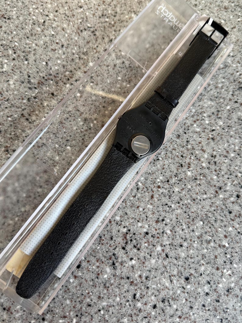 K&ouml;nnte beinhalten: Eine schwarze Swatch-Uhr mit strukturiertem Lederarmband wird in einer transparenten Kunststoffbox pr&auml;sentiert. Das Zifferblatt der Uhr ist schwarz und das Armband wird mit einer silbernen Schnalle befestigt. Die Uhr ist neu und ungetragen.