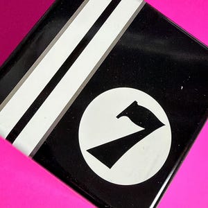 May include: Black, square tin with white stripes and a white circle containing a black graphic of a stylized number seven. The tin is set against a bright pink background.