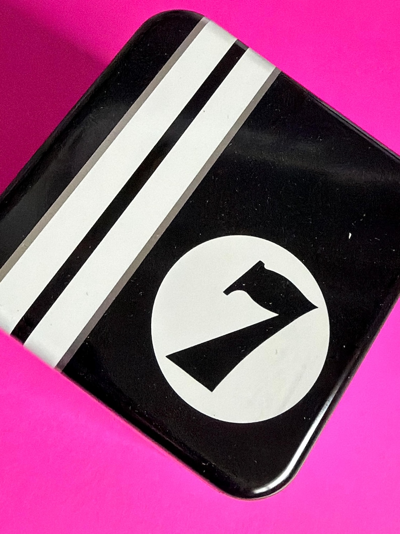 May include: Black, square tin with white stripes and a white circle containing a black graphic of a stylized number seven. The tin is set against a bright pink background.