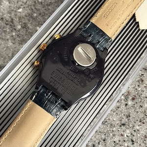 May include: Black Swatch watch with a gold-tone bezel and a black leather strap. The watch face is black with white markings. The back of the watch has the text "WATERRESISTANT", "PATENTED", "SWISS MADE", "TWENTY TWO (22)", "JEWELS 26", "BATTERY", "Swatch", "genuine leather", and "Made in Switzerland".