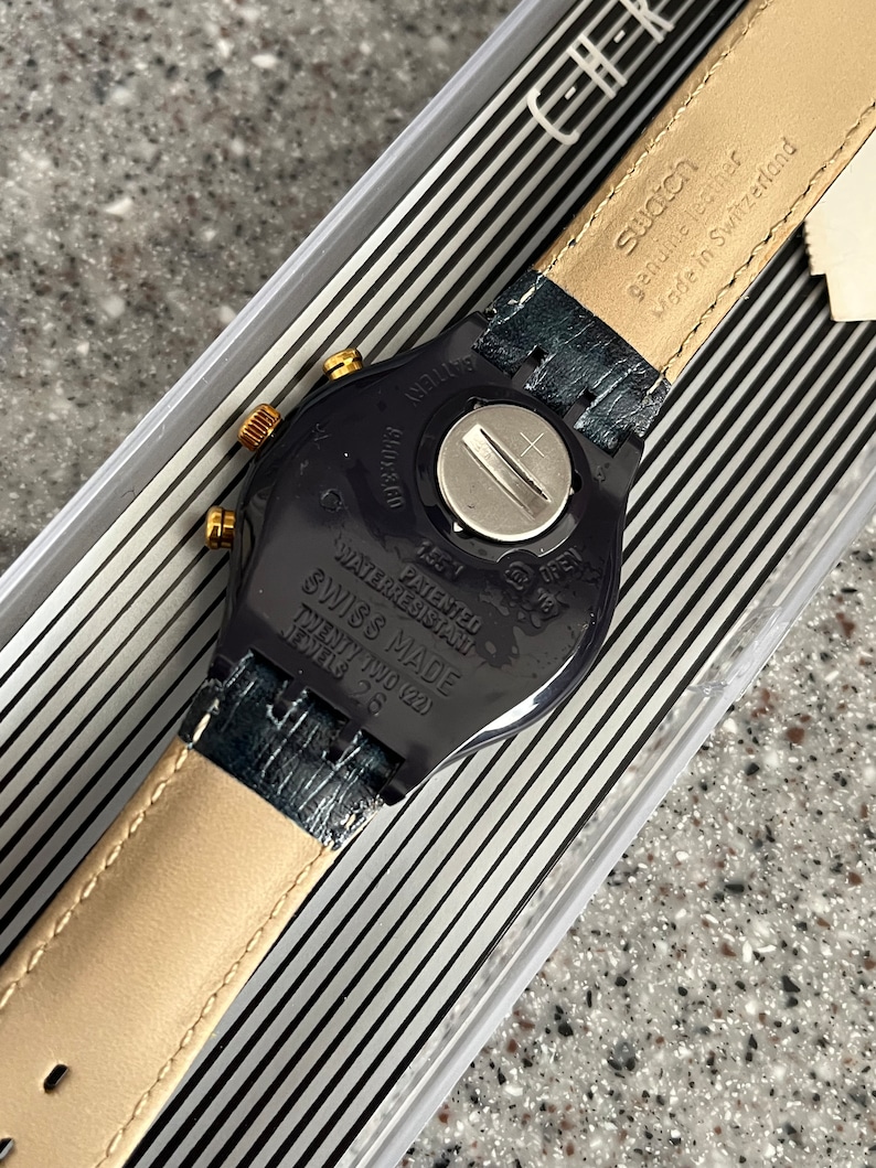 May include: Black Swatch watch with a gold-tone bezel and a black leather strap. The watch face is black with white markings. The back of the watch has the text "WATERRESISTANT", "PATENTED", "SWISS MADE", "TWENTY TWO (22)", "JEWELS 26", "BATTERY", "Swatch", "genuine leather", and "Made in Switzerland".