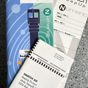 May include: Swatch BeepUp product packaging, featuring the brand's logo and the word "Numeric." The packaging includes a blue and white booklet, along with several white documents. The address of Swatch AG, BeepUp Division, is visible.