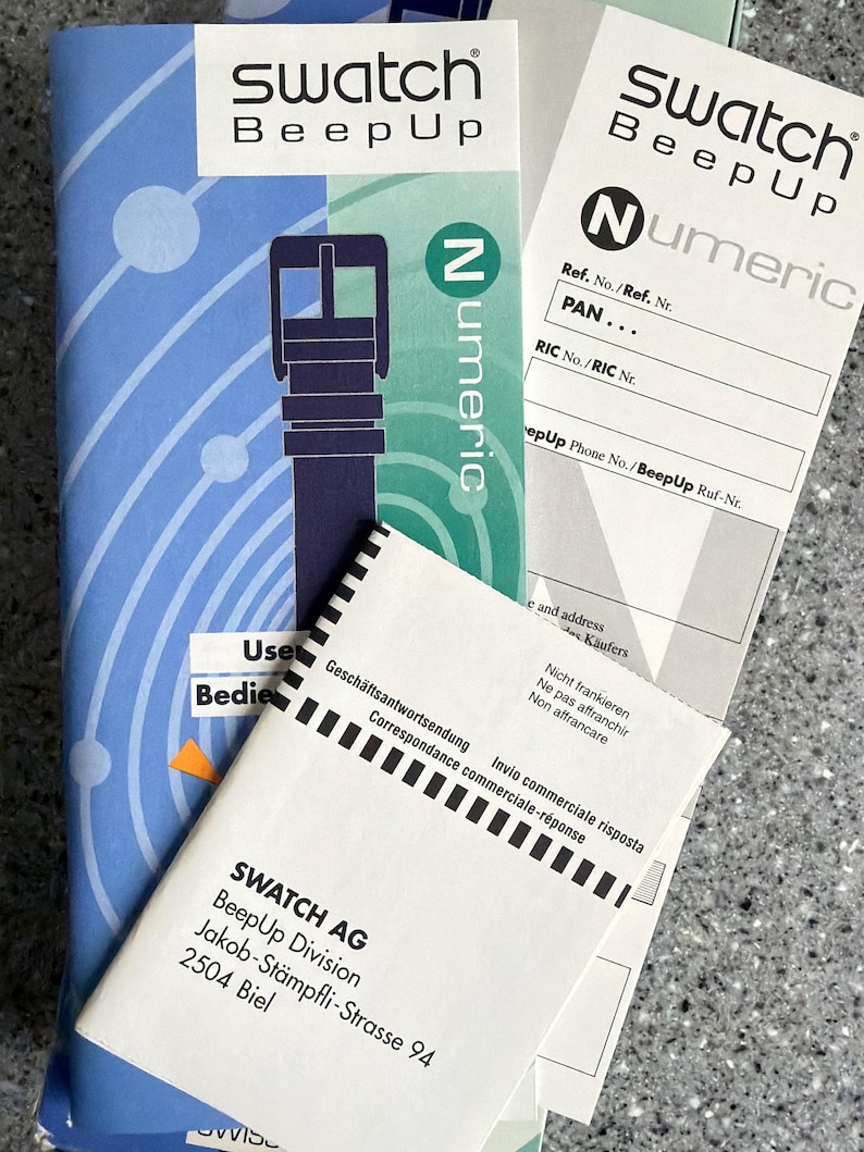 May include: Swatch BeepUp product packaging, featuring the brand's logo and the word "Numeric." The packaging includes a blue and white booklet, along with several white documents. The address of Swatch AG, BeepUp Division, is visible.