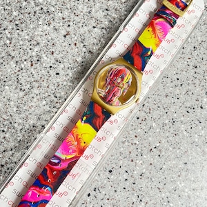 May include: A Swatch Club watch with a gold-tone case and a colorful, abstract design on the face and strap. The strap features a vibrant mix of red, yellow, blue, and pink. The watch is in its original packaging.