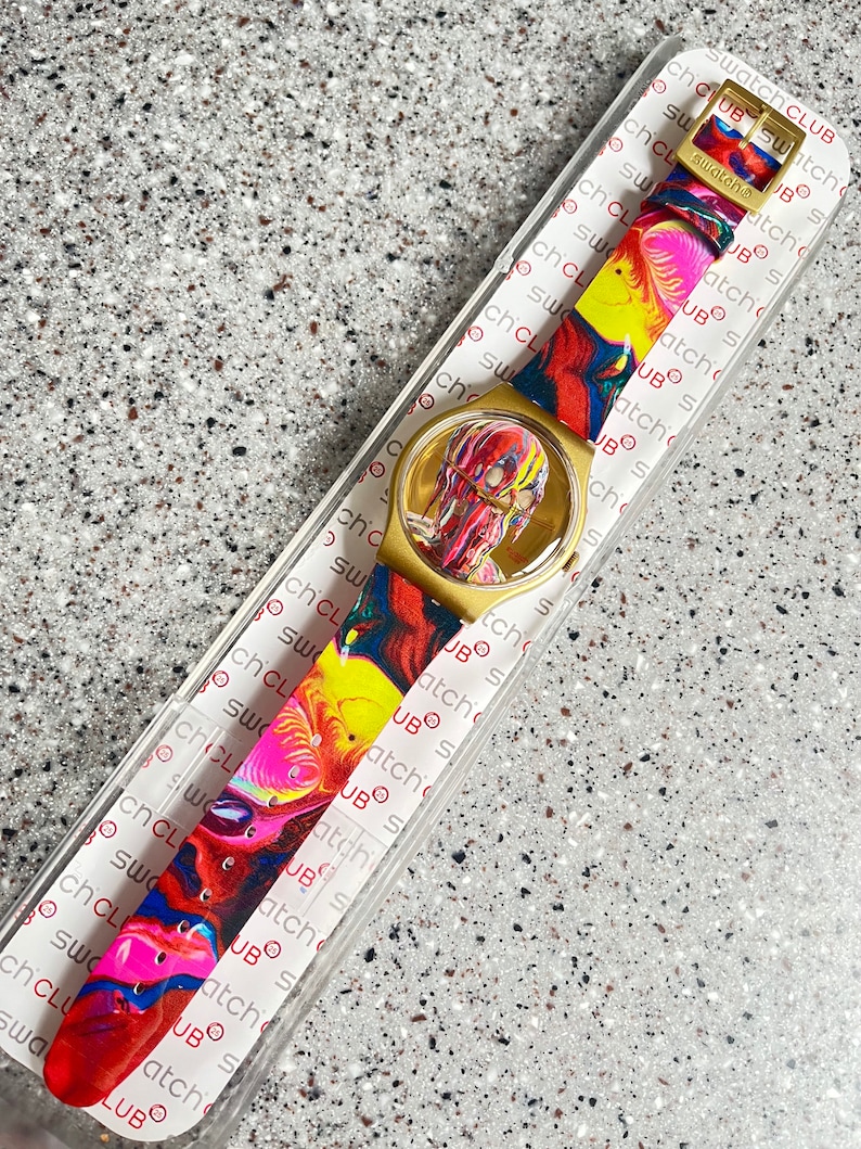 May include: A Swatch Club watch with a gold-tone case and a colorful, abstract design on the face and strap. The strap features a vibrant mix of red, yellow, blue, and pink. The watch is in its original packaging.