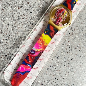 May include: A gold-toned Swatch watch with a colorful, abstract design on the band and face. The watch is in its original packaging, with the brand name visible. The band features a mix of red, yellow, blue, and pink hues.