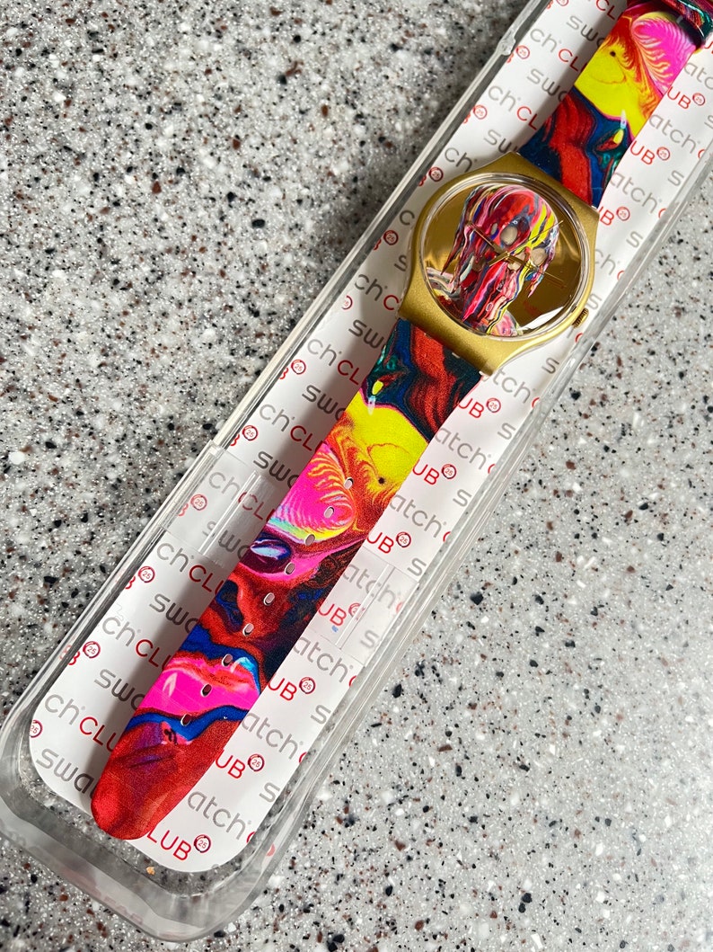 May include: A gold-toned Swatch watch with a colorful, abstract design on the band and face. The watch is in its original packaging, with the brand name visible. The band features a mix of red, yellow, blue, and pink hues.