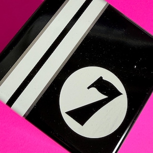 May include: Black square tin with white stripes and a white circle containing a black number seven graphic. The tin is set against a bright pink background.