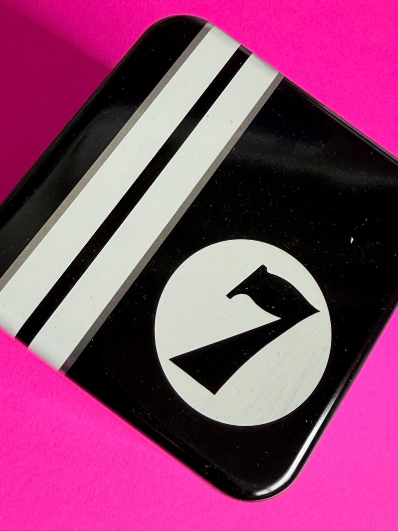 May include: Black square tin with white stripes and a white circle containing a black number seven graphic. The tin is set against a bright pink background.