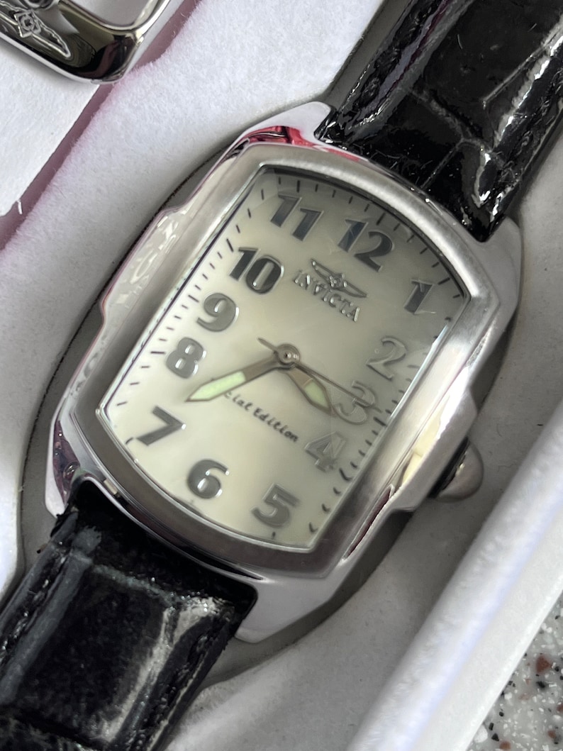 Puede incluir: Un reloj plateado con una esfera blanca y una correa de cuero negra. La esfera del reloj tiene marcadores de hora en n&uacute;meros romanos negros y una aguja de segundos plateada. La esfera del reloj tambi&eacute;n tiene el texto "Invicta" y "Flat Edition".