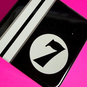 May include: Black, square tin with white stripes and a white circle containing a black number seven. The tin sits on a bright pink surface. The design is simple and graphic.