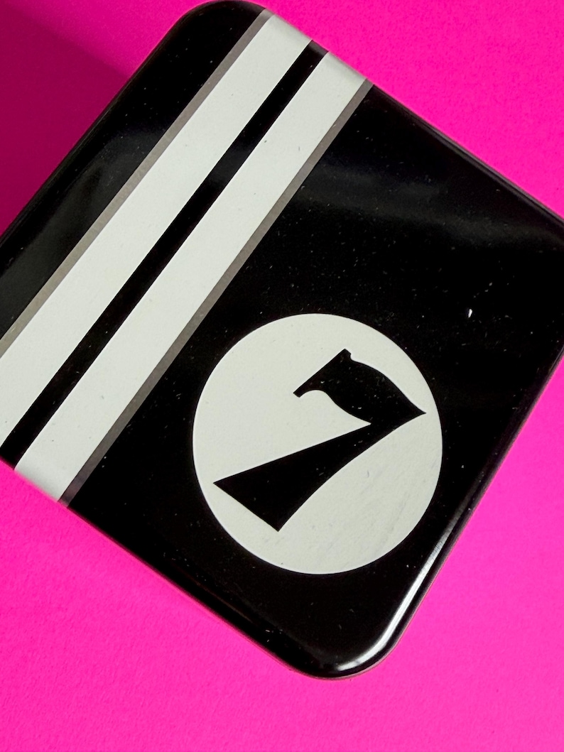 May include: Black, square tin with white stripes and a white circle containing a black number seven. The tin sits on a bright pink surface. The design is simple and graphic.