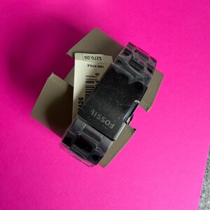 May include: A Fossil brand watch with a silver band and a black face, presented in a cardboard box. The watch is partially visible, with the brand name prominently displayed. The background is a bright pink surface.