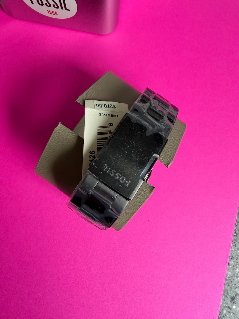 May include: A Fossil brand watch with a silver band and a black face, presented in a cardboard box. The watch is partially visible, with the brand name prominently displayed. The background is a bright pink surface.