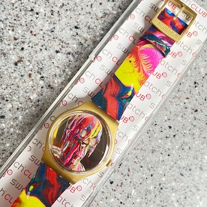 May include: A Swatch watch with a gold-colored case and a colorful, abstract design on the face and strap. The strap features a gold-colored buckle. The watch is in its original packaging.