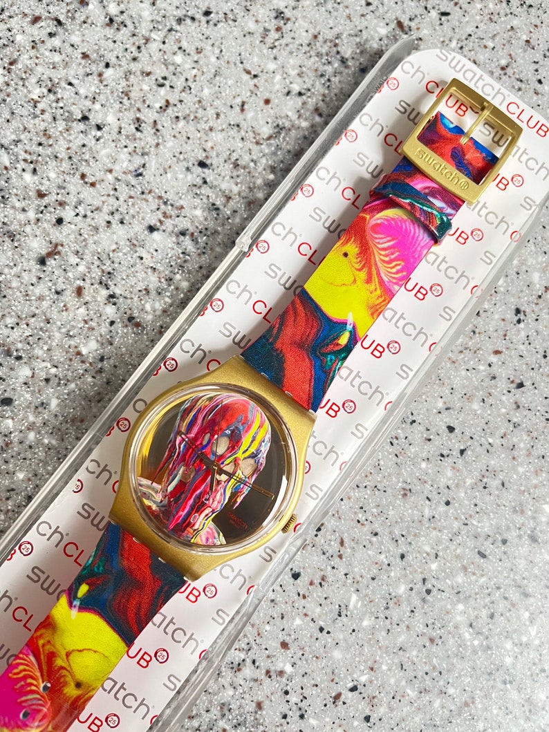 May include: A Swatch watch with a gold-colored case and a colorful, abstract design on the face and strap. The strap features a gold-colored buckle. The watch is in its original packaging.