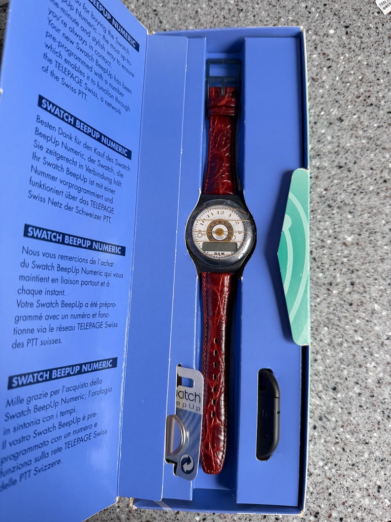 May include: A vintage Swatch BeepUp Numeric watch with a red leather band is displayed in its original blue box. The watch face is white with black numerals and a gold-colored center. A black accessory is also included.