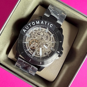 May include: Black automatic watch with a skeleton dial, visible gears, and a black metal band. The watch face has white hands and markers. The word "AUTOMATIC" is printed in white on the top of the watch face. The watch is in a beige box.