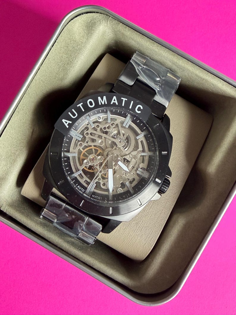 May include: Black automatic watch with a skeleton dial, visible gears, and a black metal band. The watch face has white hands and markers. The word "AUTOMATIC" is printed in white on the top of the watch face. The watch is in a beige box.