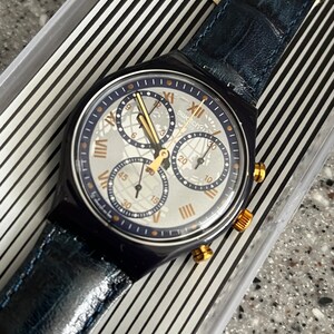 May include: A blue and silver Swatch chronograph watch with a blue leather band. The watch face has a white background with black Roman numeral hour markers and gold accents.