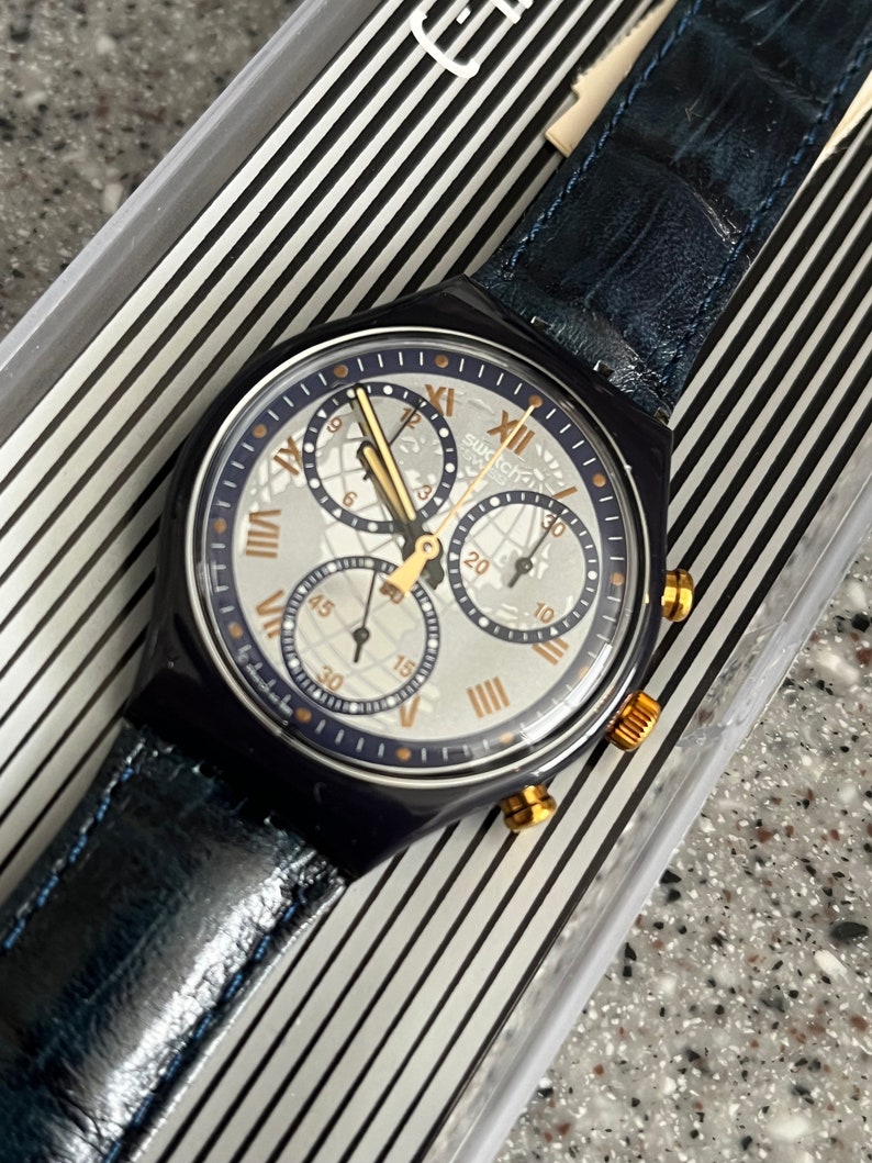 May include: A blue and silver Swatch chronograph watch with a blue leather band. The watch face has a white background with black Roman numeral hour markers and gold accents.