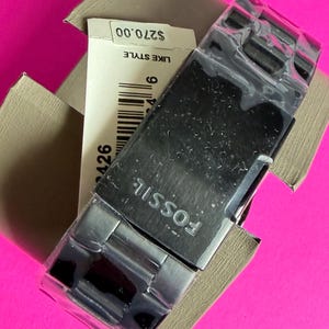 May include: A new Fossil watch with a silver-tone stainless steel band and a black watch face, still in its original packaging. The watch is displayed on a bright pink background. The watch's brand name, "FOSSIL", is visible on the watch face.