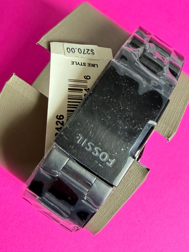 May include: A new Fossil watch with a silver-tone stainless steel band and a black watch face, still in its original packaging. The watch is displayed on a bright pink background. The watch's brand name, "FOSSIL", is visible on the watch face.
