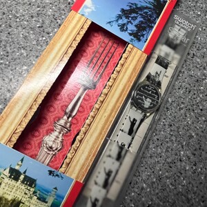 Op de afbeelding: Een zilveren vork in een rood en gouden frame met een witte achtergrond. De vork zit in een doos met een wit horloge met een zwart-witte afbeelding van een kasteel op de wijzerplaat. De doos heeft de tekst "Swatch Quartz" en "Swiss Made" erop gedrukt.