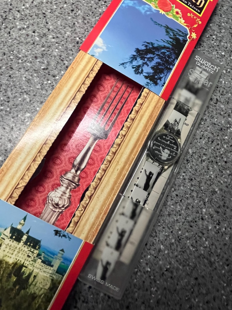 Op de afbeelding: Een zilveren vork in een rood en gouden frame met een witte achtergrond. De vork zit in een doos met een wit horloge met een zwart-witte afbeelding van een kasteel op de wijzerplaat. De doos heeft de tekst "Swatch Quartz" en "Swiss Made" erop gedrukt.