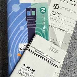 May include: A collection of Swatch BeepUp product packaging and paperwork. The packaging features a blue and white design with a watch strap illustration and the word "Numeric". Several white documents with text are also visible.