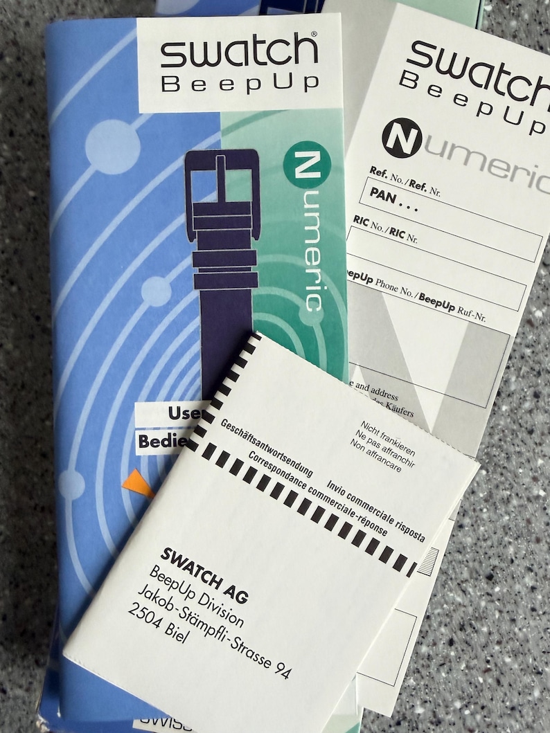 May include: A collection of Swatch BeepUp product packaging and paperwork. The packaging features a blue and white design with a watch strap illustration and the word "Numeric". Several white documents with text are also visible.