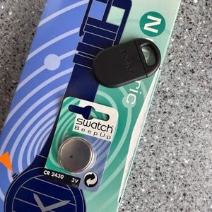 May include: A black Swatch watch accessory and a watch battery are displayed on a patterned background. The battery is in a green and white package with the text "Swatch Beep Up". The accessory is oval-shaped with a loop.