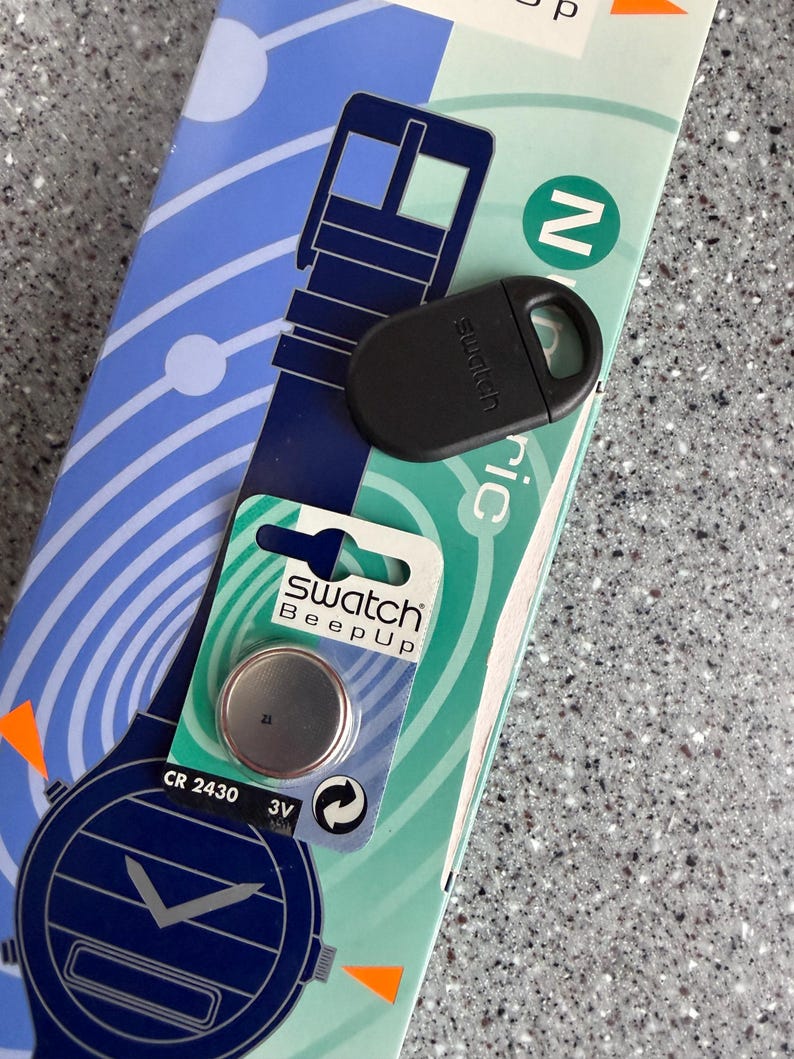 May include: A black Swatch watch accessory and a watch battery are displayed on a patterned background. The battery is in a green and white package with the text "Swatch Beep Up". The accessory is oval-shaped with a loop.