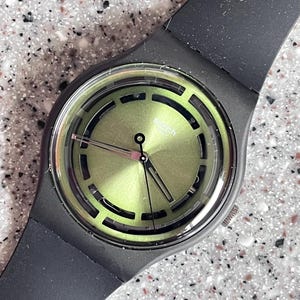 May include: Black wristwatch with a green face and black strap. The watch face features a circular design with cut-out shapes. The watch hands are black. The watch is set against a speckled background.
