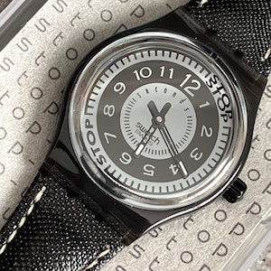 Puede incluir: Un reloj de pulsera negro con una esfera blanca y agujas negras. El reloj tiene una correa de cuero negro con costuras blancas. La esfera del reloj tiene las palabras "Stop" y "Seconds" impresas en ella.