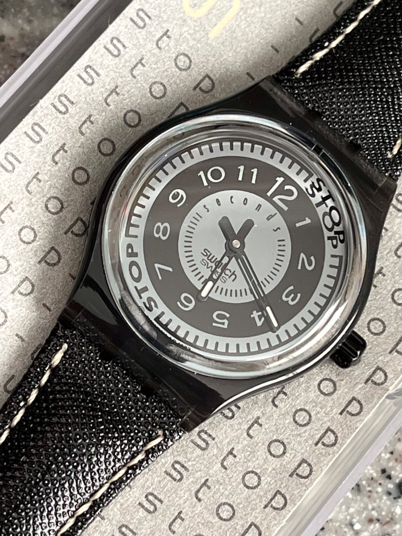 Puede incluir: Un reloj de pulsera negro con una esfera blanca y agujas negras. El reloj tiene una correa de cuero negro con costuras blancas. La esfera del reloj tiene las palabras "Stop" y "Seconds" impresas en ella.