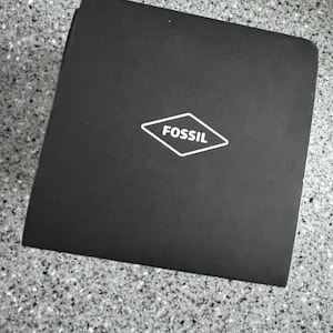 May include: A black square box with the brand name "FOSSIL" in a white diamond-shaped logo. The box is on a speckled gray surface.